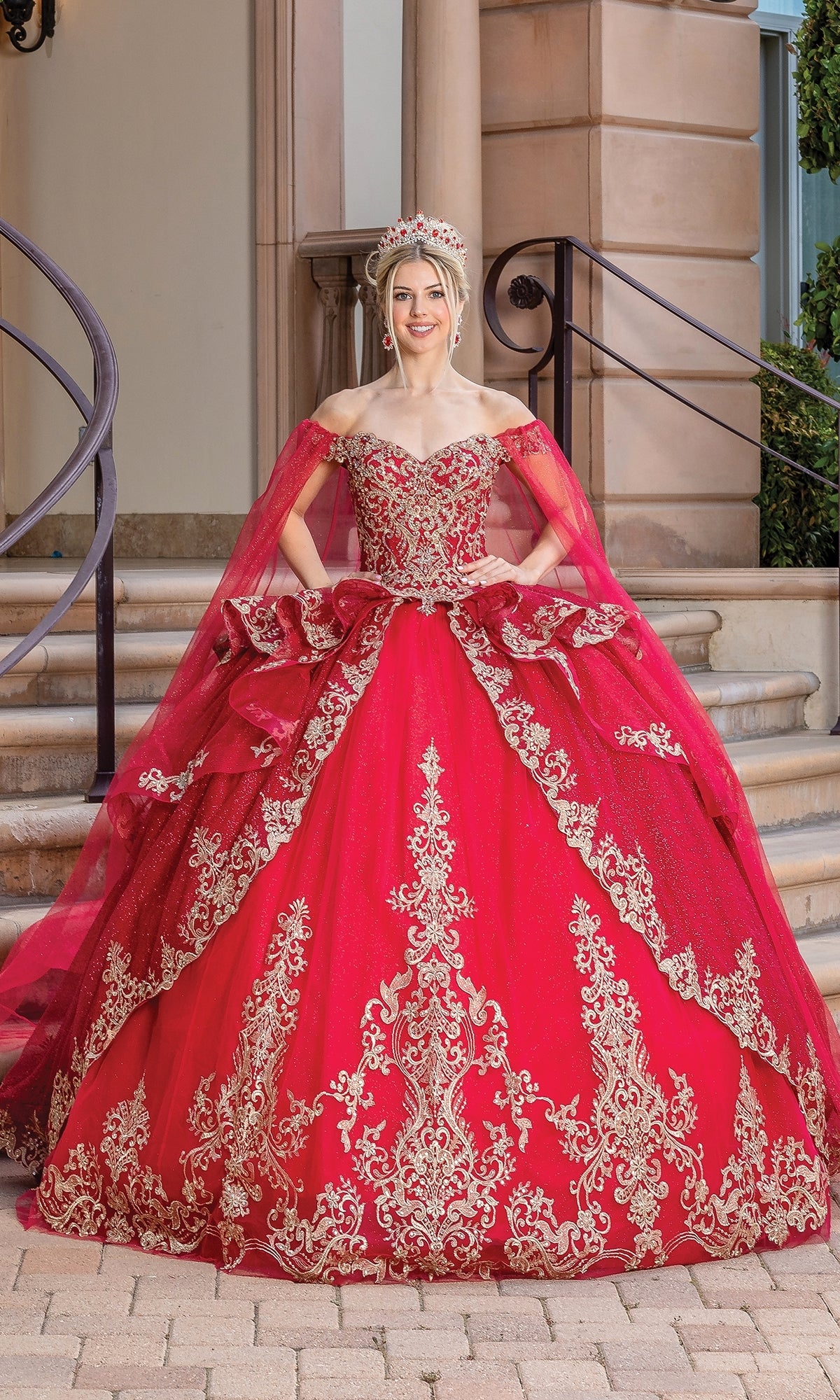 A woman stands on steps wearing the Quinceanera Dress 1720 By Dancing Queen, an off-the-shoulder bright red ball gown with gold embroidery, a matching cape, and a jeweled crown.